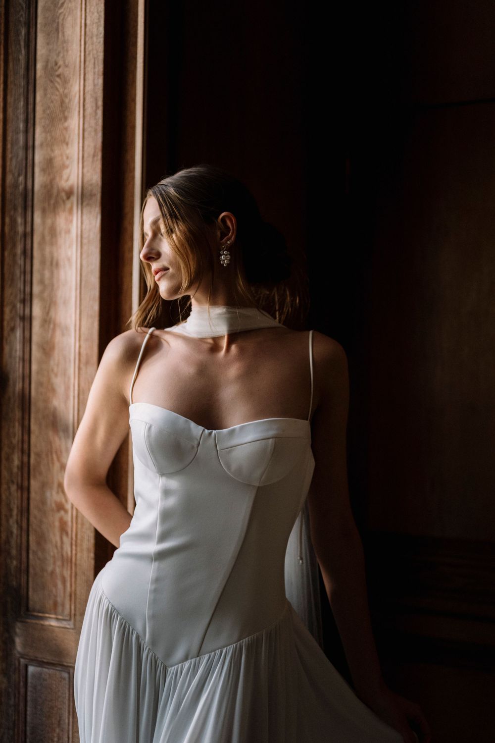 A person in a white dress stands near wooden panels in dramatic lighting creating an artistic bridal portrait.