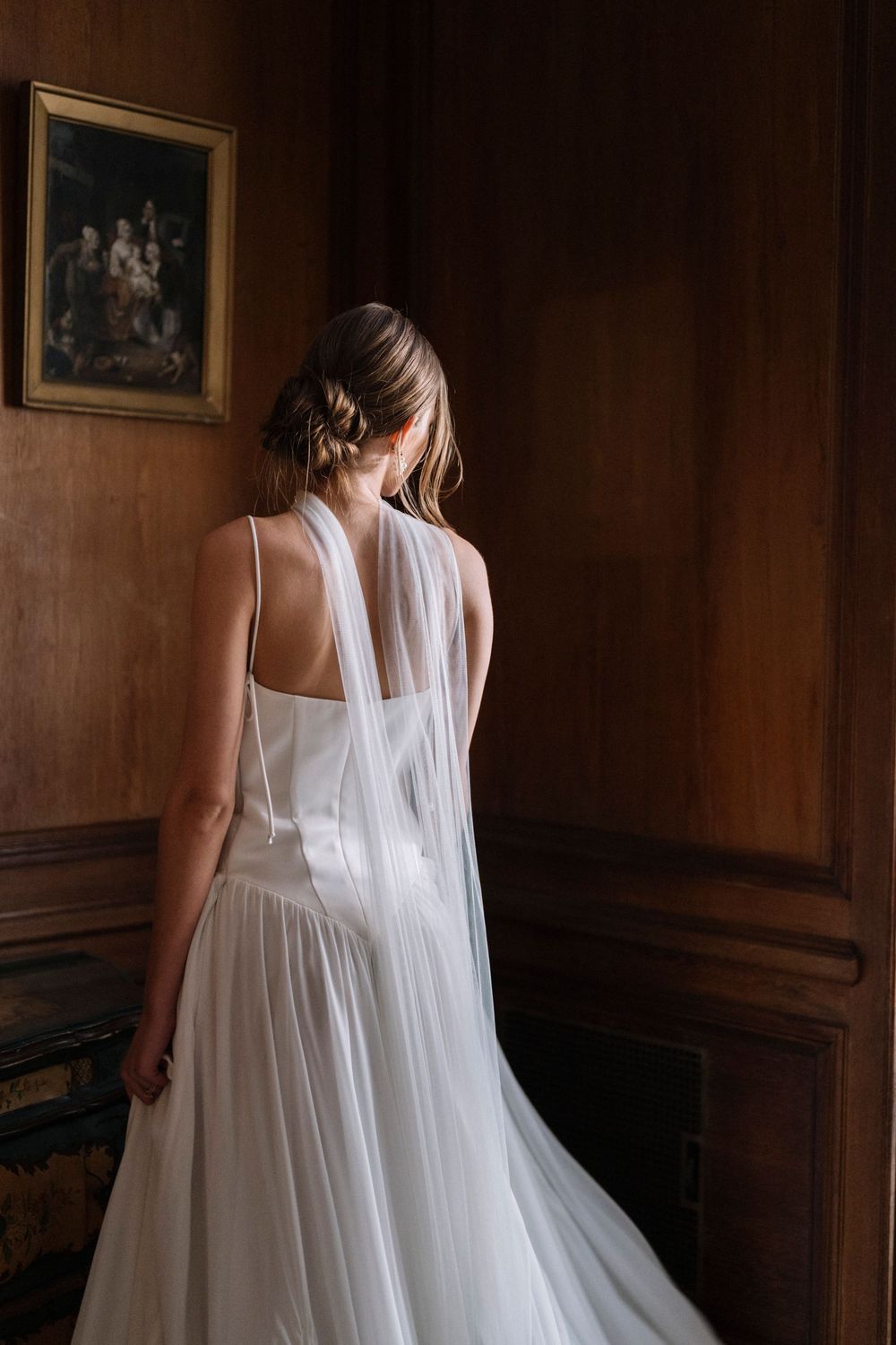 A flowing white dress with sheer fabric cascades down as someone walks through a dark wood-paneled hallway.