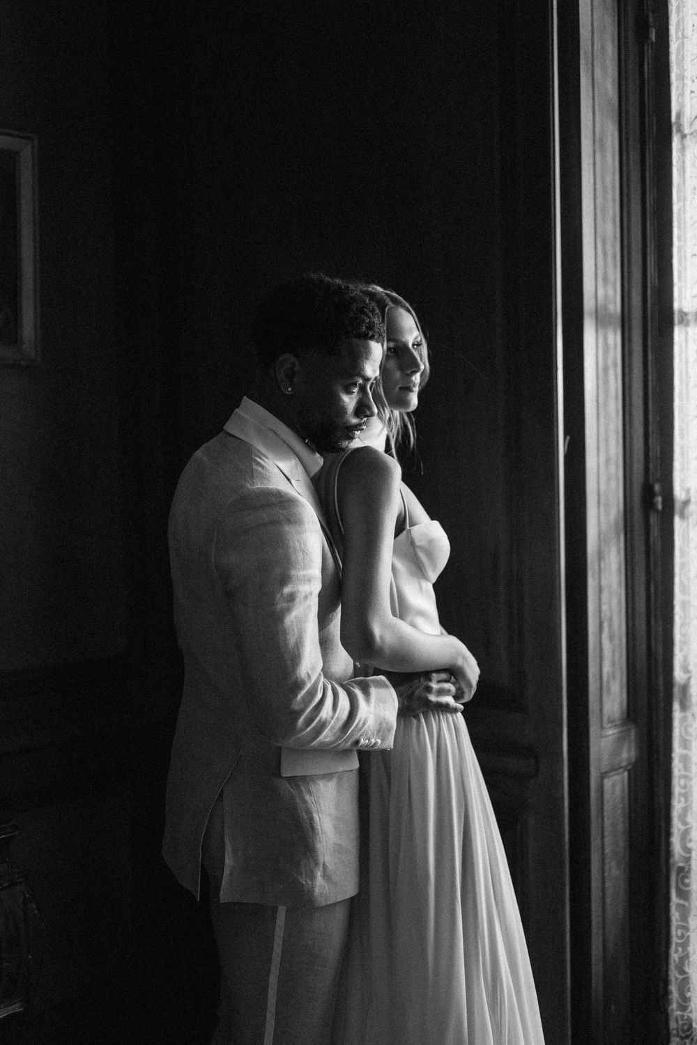 A romantic black and white wedding photo of a couple standing by a window in formal attire.