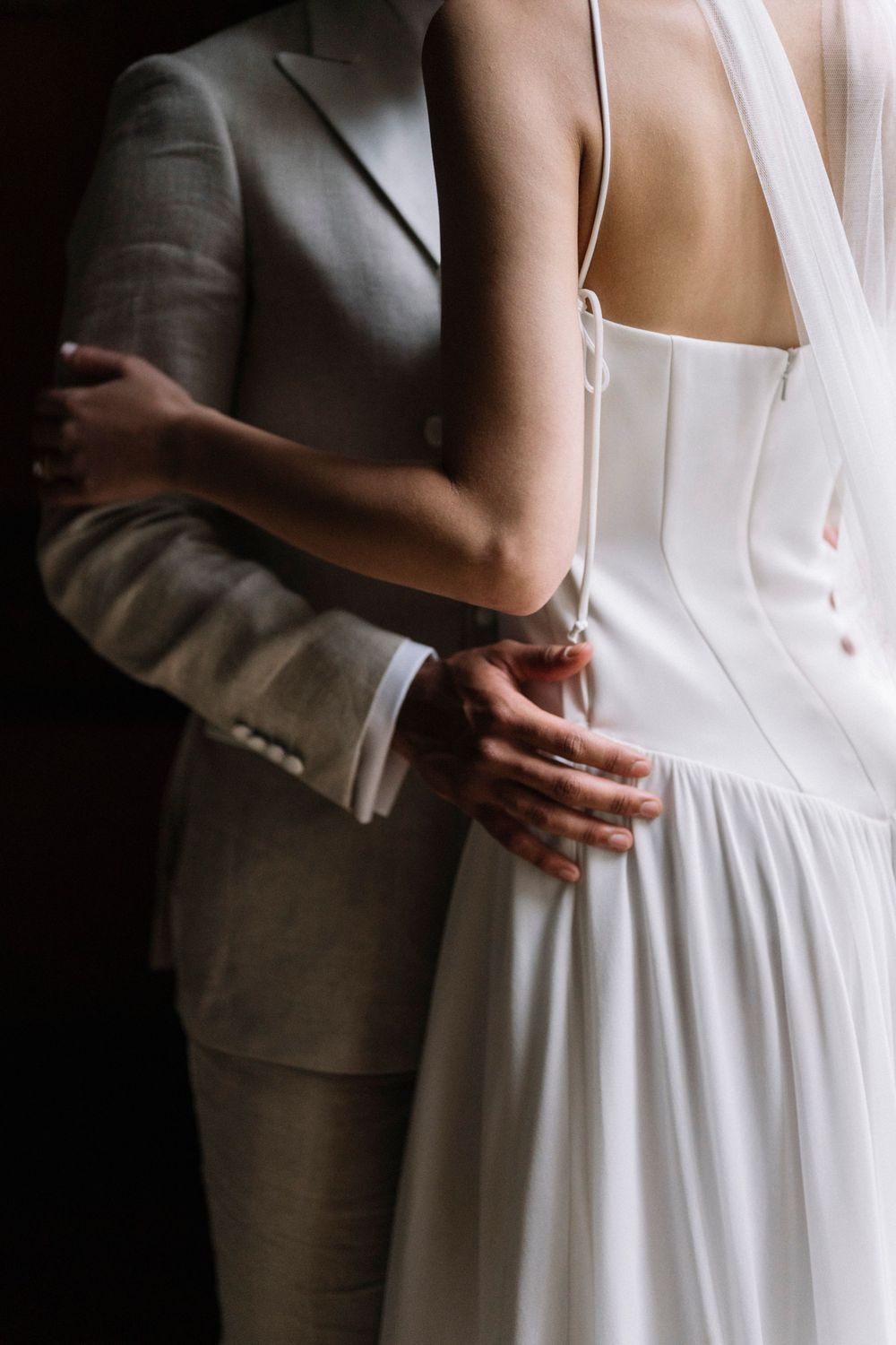 Close-up detail shot of a couple embracing, showing the back of a white wedding dress and light gray suit.