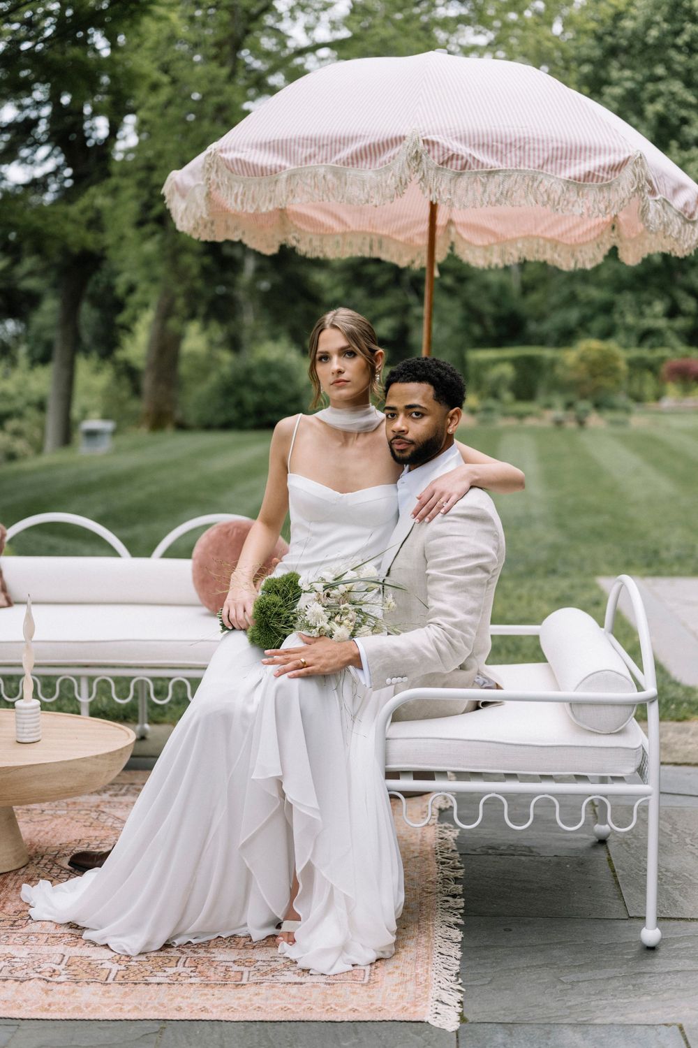 Wedding couple sitting together under a vintage parasol on outdoor patio furniture.