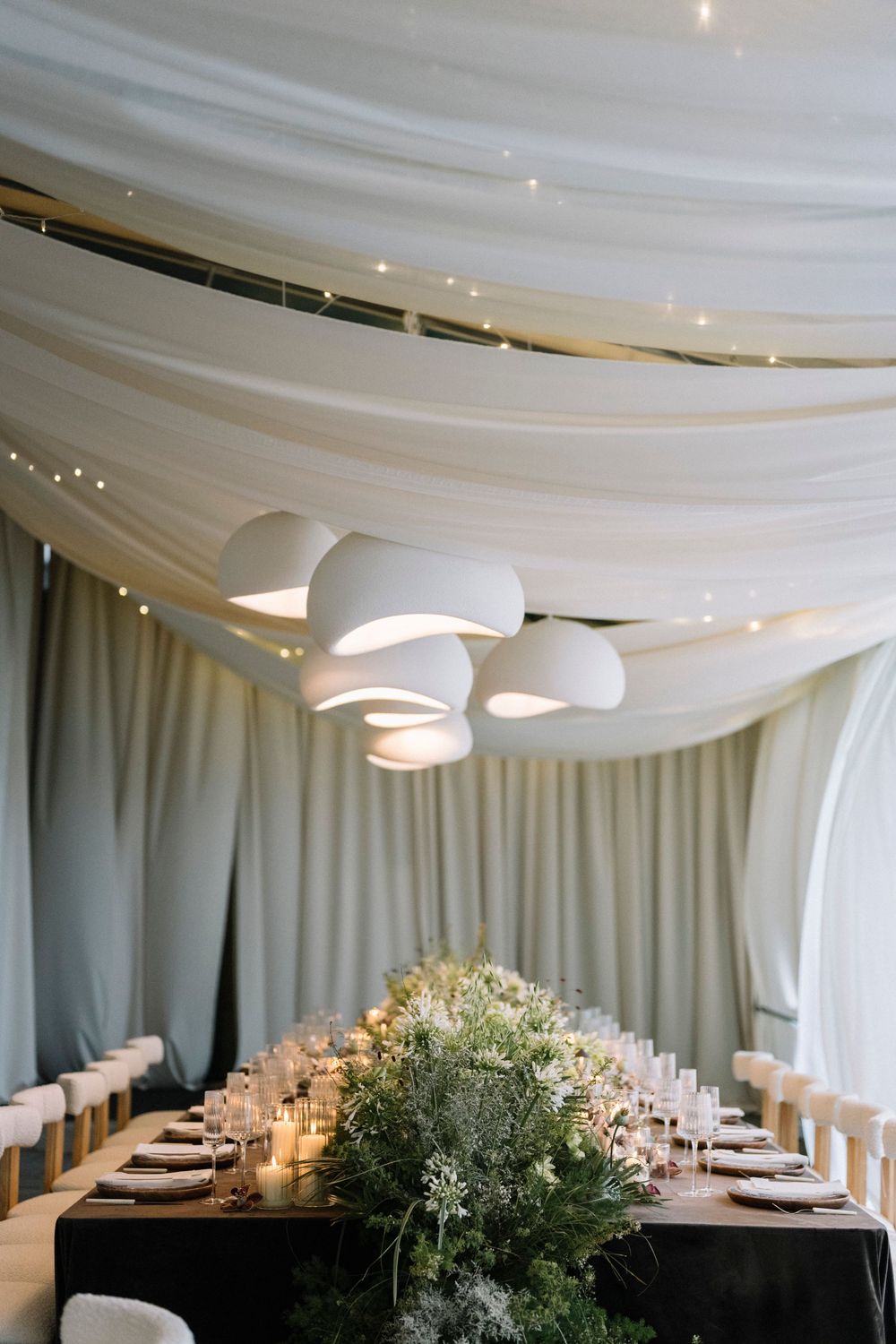 Modern wedding reception venue with white draped ceiling, pendant lights, and elegant table settings decorated with greenery.