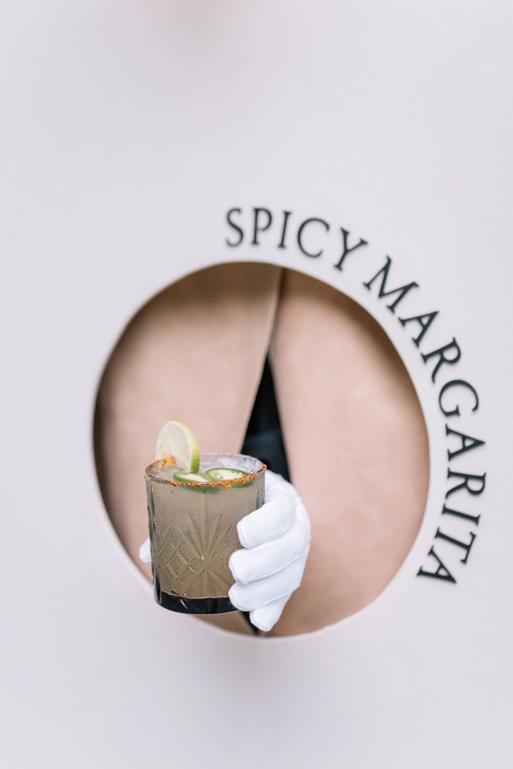 A hand in a white glove holds a spicy margarita cocktail viewed through a circular window with text above.