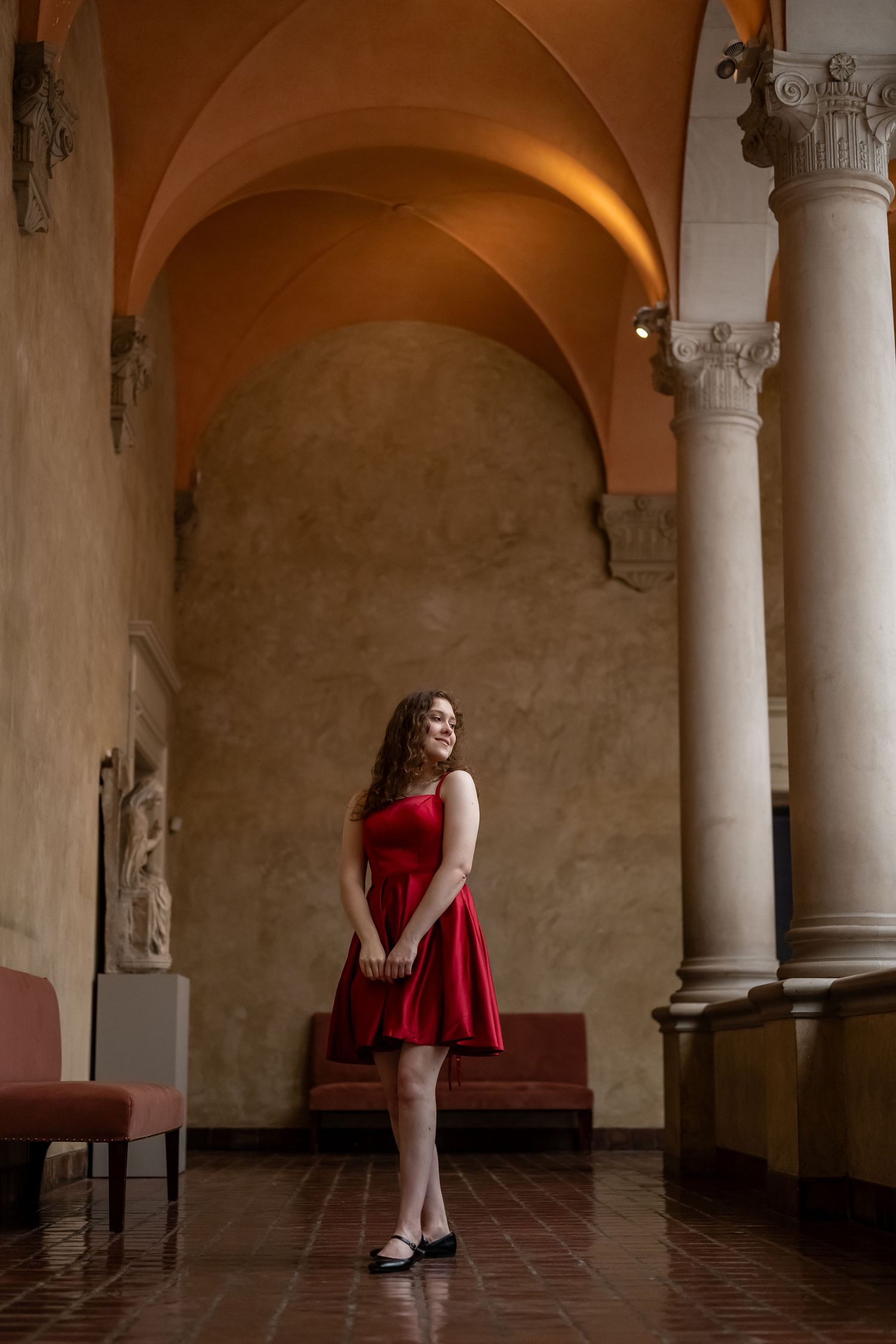 A woman in a red dress sits contemplatively in an elegant arched corridor with classical columns and warm lighting.