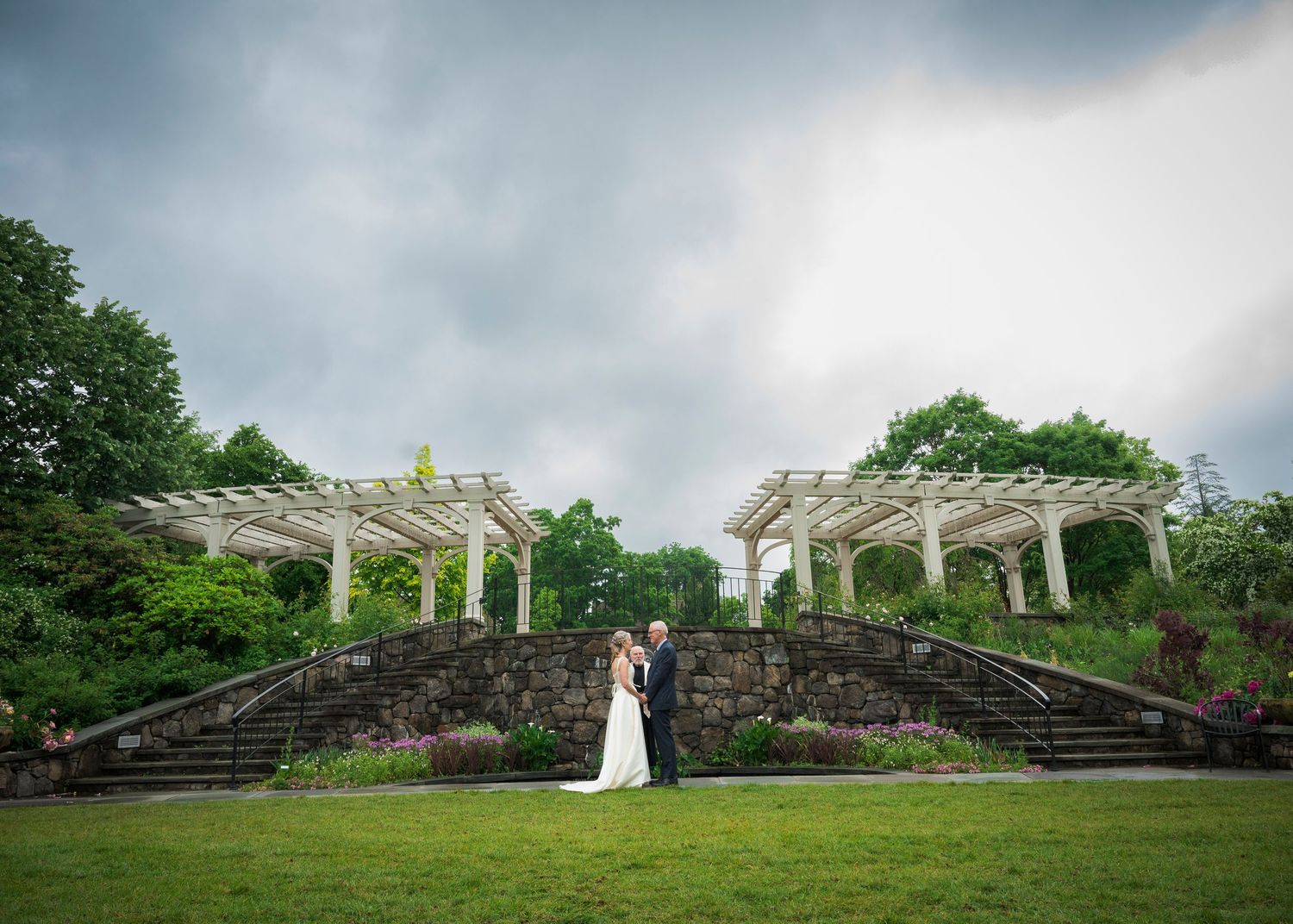 A couple stands on a stone staircase with white pergolas and dramatic storm clouds in an elegant garden setting.
