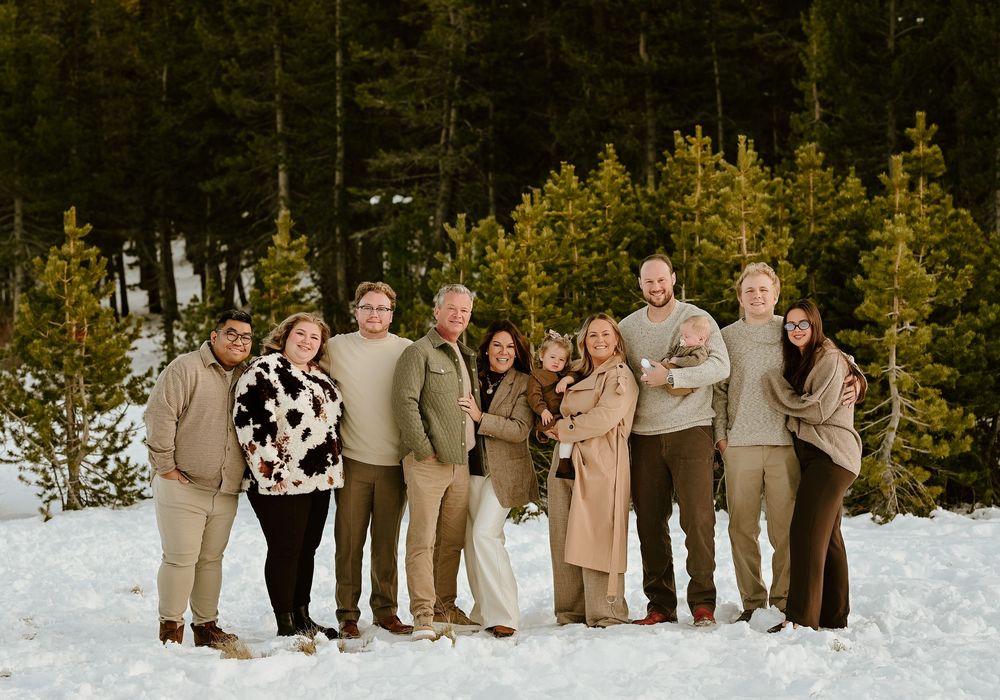A wedding party in neutral tones poses together in a snowy winter landscape with evergreen trees in the background.