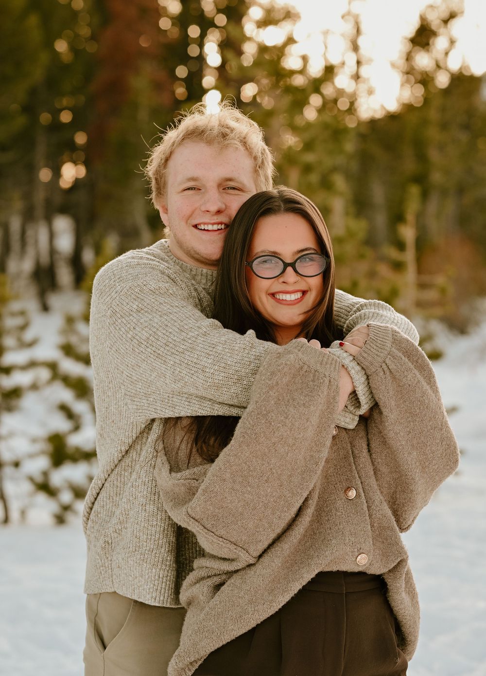 A couple in cozy beige sweaters embrace while standing in a snowy winter forest with warm sunset lighting in the background.