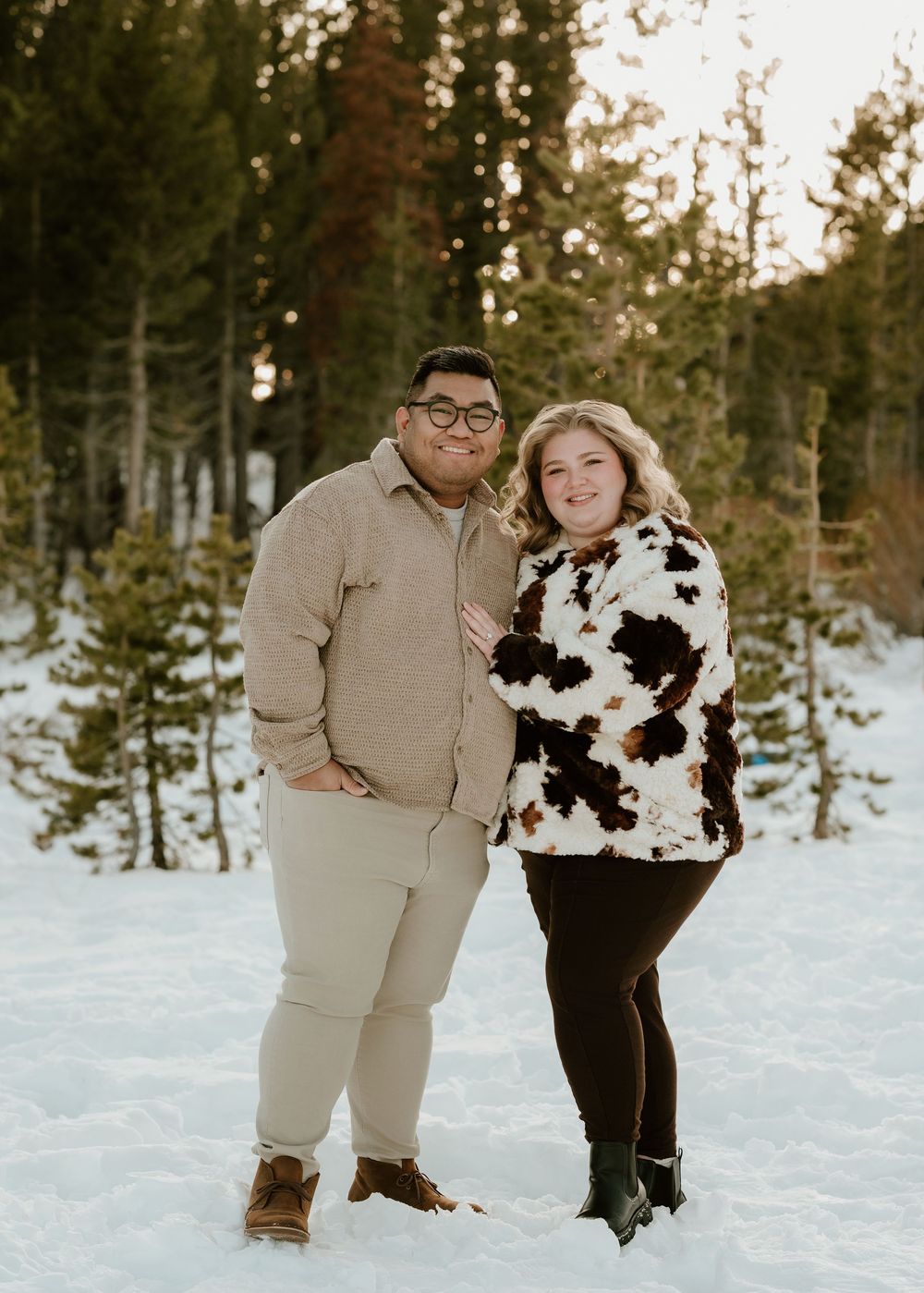A couple shares a joyful moment together in a snowy winter forest setting with pine trees in the background.