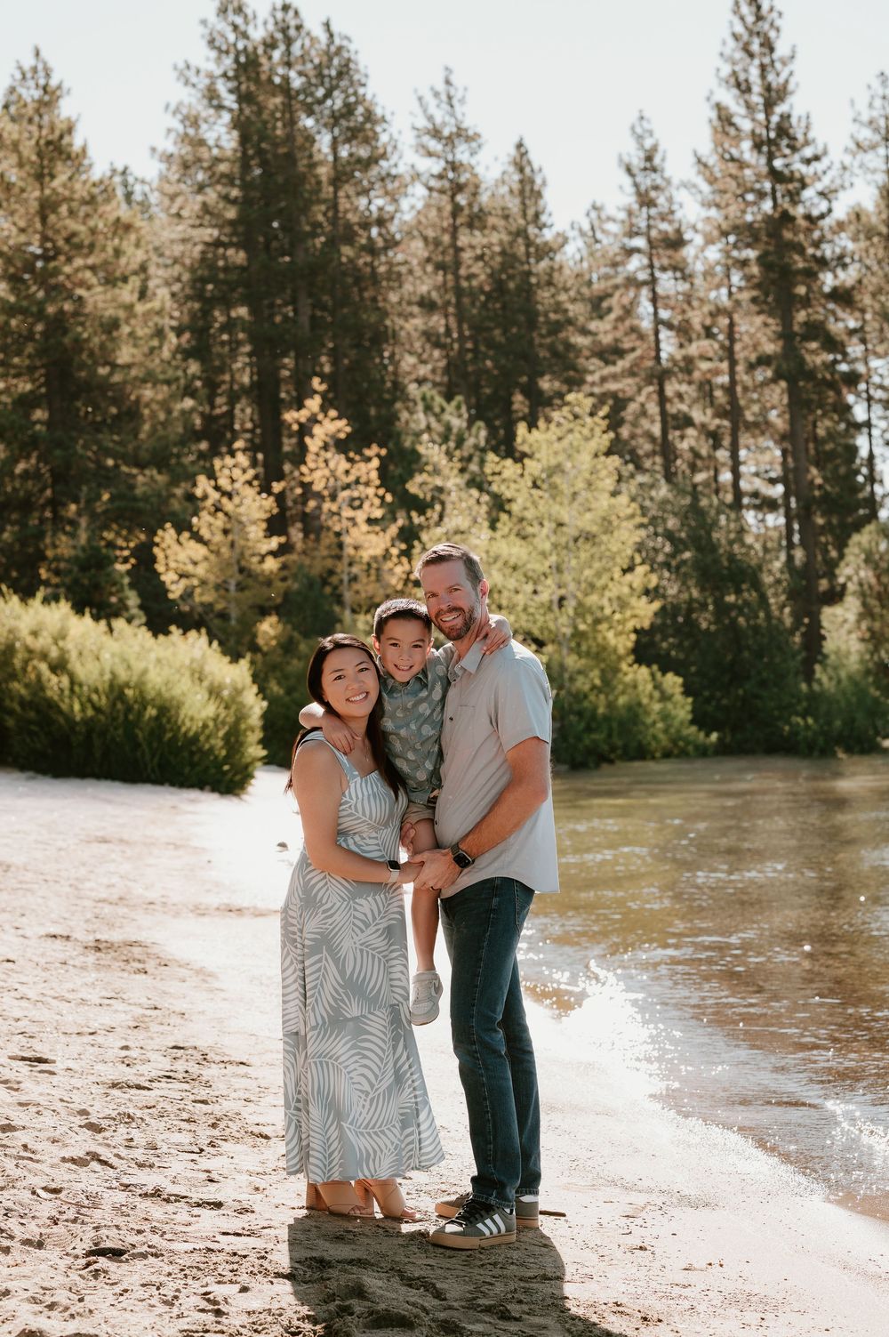 A family stands together on a beach by a lake surrounded by tall pine trees on a sunny afternoon.