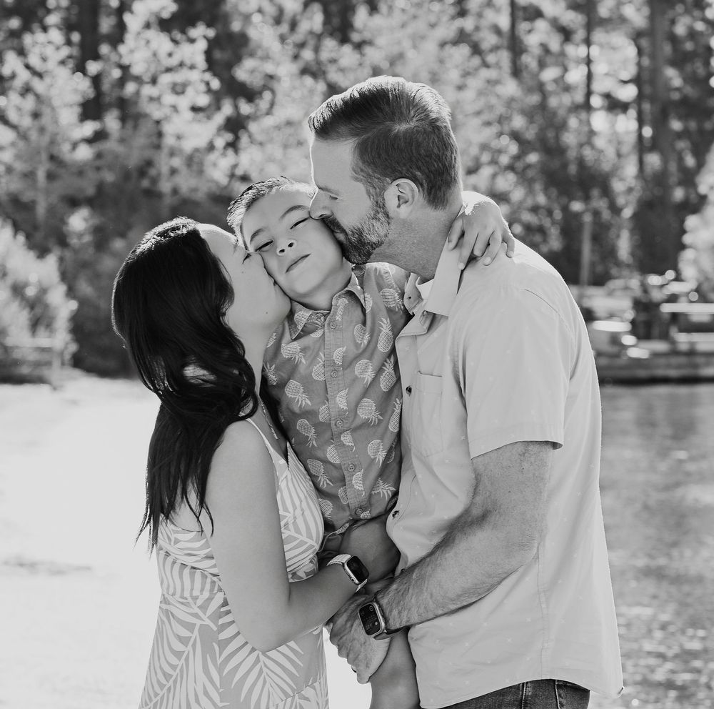 A tender family moment captured in black and white photography outdoors with trees in the background.