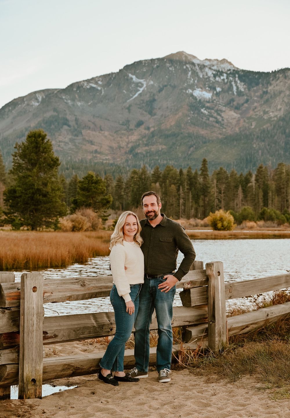 Couple sits together on rustic wooden fence with snow-capped mountains and evergreen forest in background at sunset.