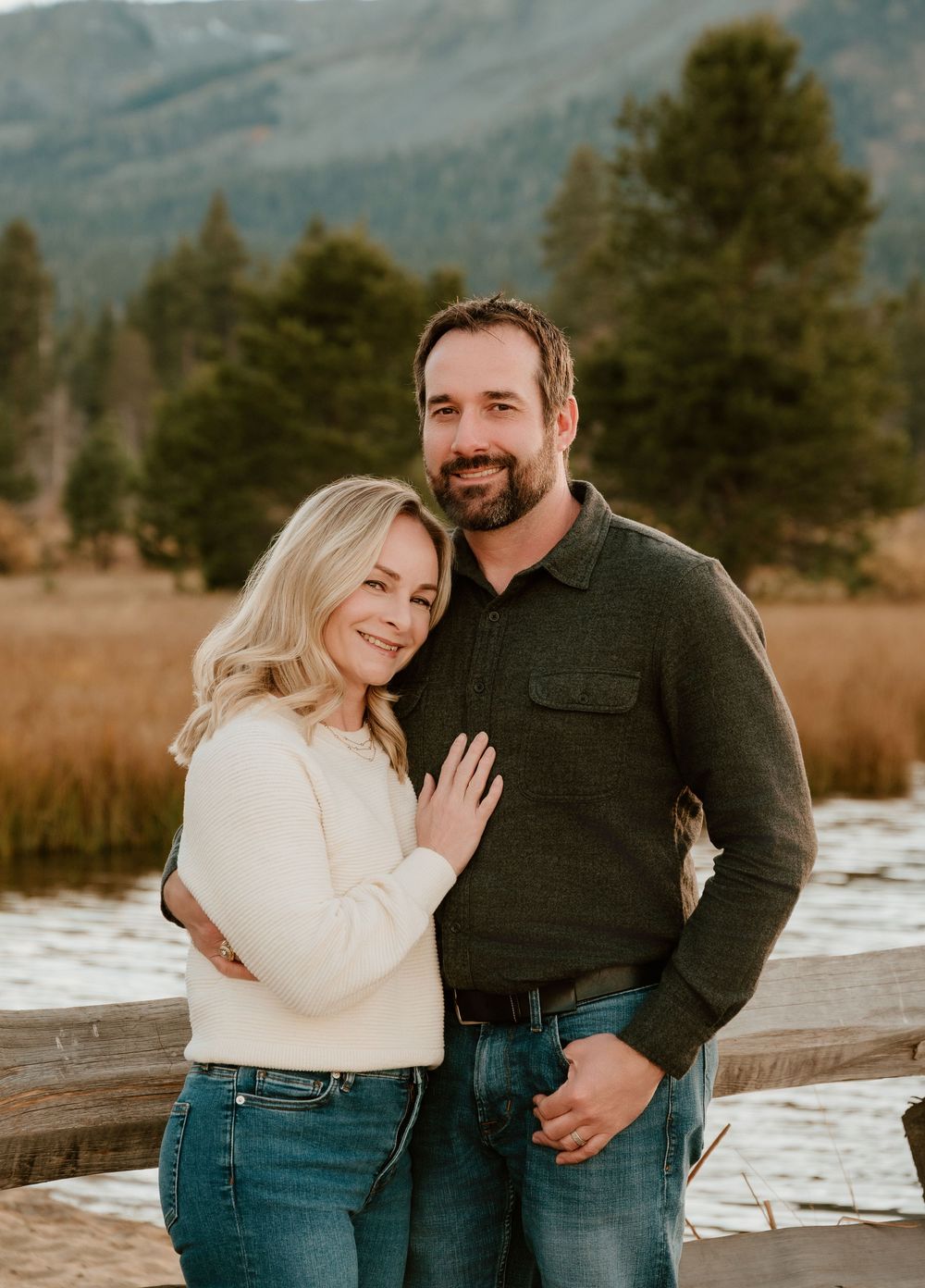 A couple poses together outdoors in warm sweaters with mountains and pine trees in the background on a fall day.