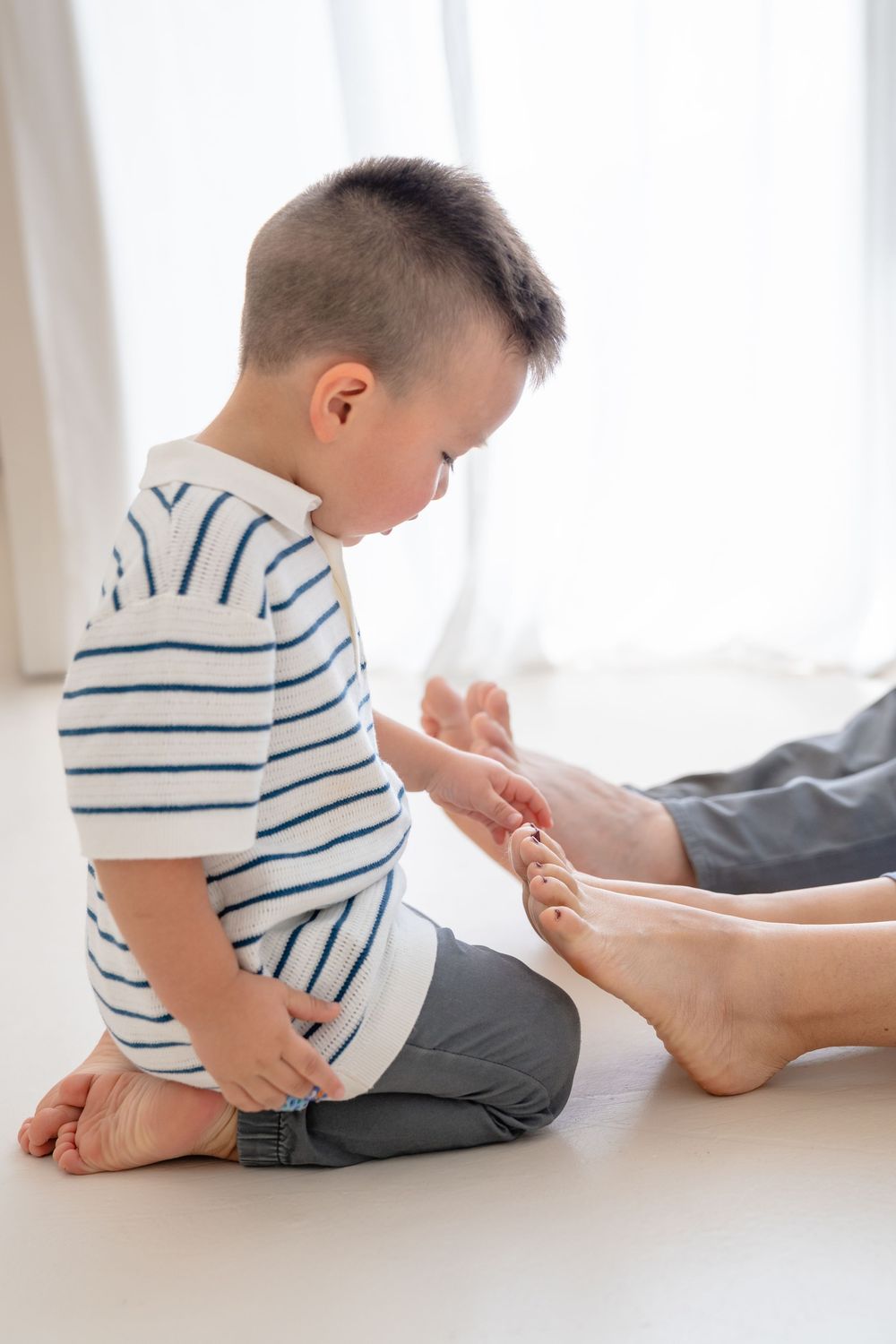 A child in a striped shirt kneels on a white surface while examining bare feet nearby.