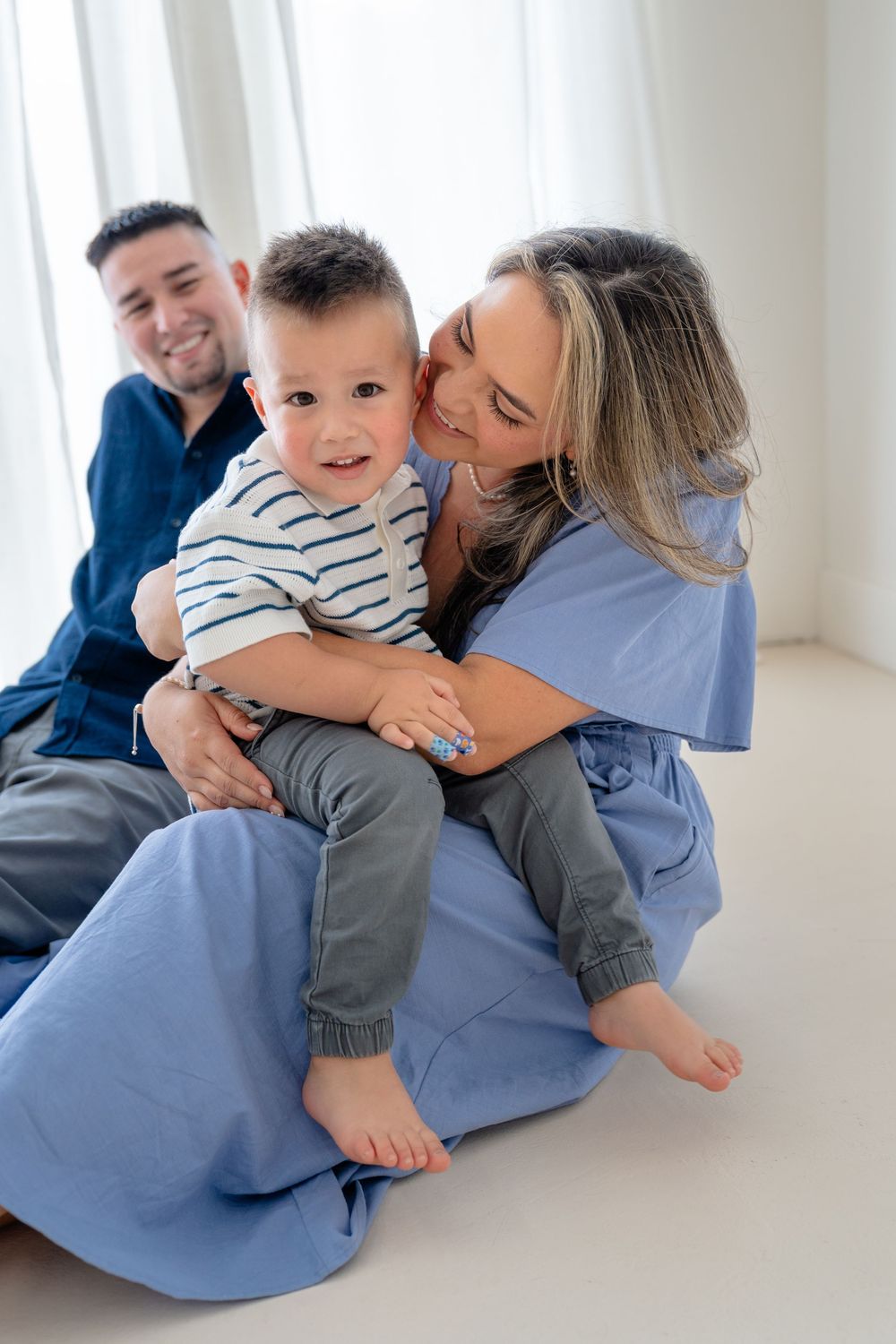 Family shares playful moments together on white floor with parent wearing blue scrubs and child in striped shirt.