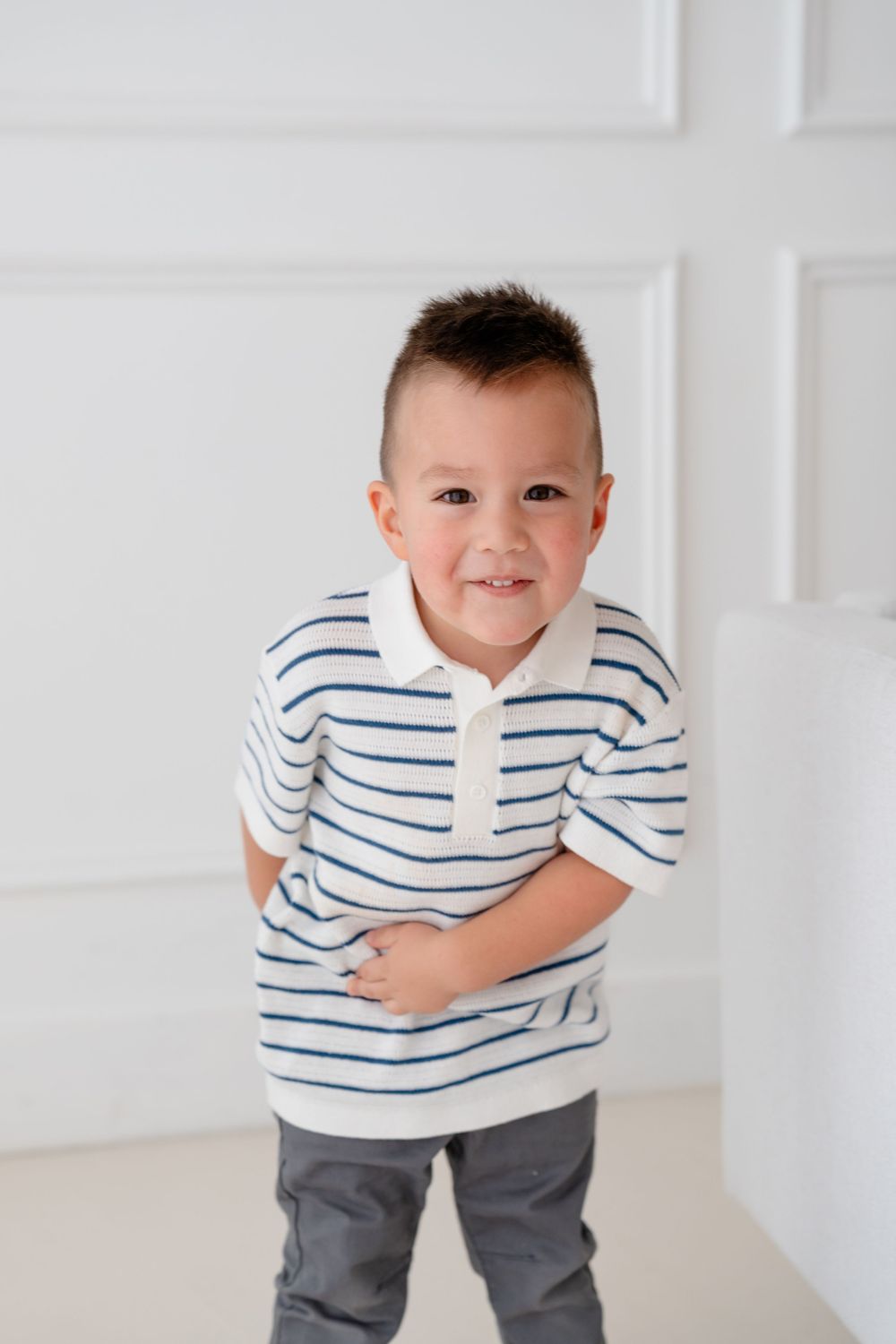 Young child in a white and blue striped shirt poses playfully against a white wall.