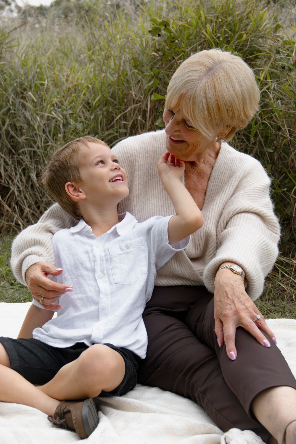 Grandmother and young grandson share a joyful moment sitting together outdoors on a blanket surrounded by green grass.