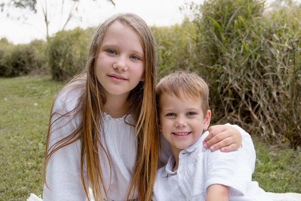 Two smiling children in white shirts sitting outdoors on grass with green foliage in the background.