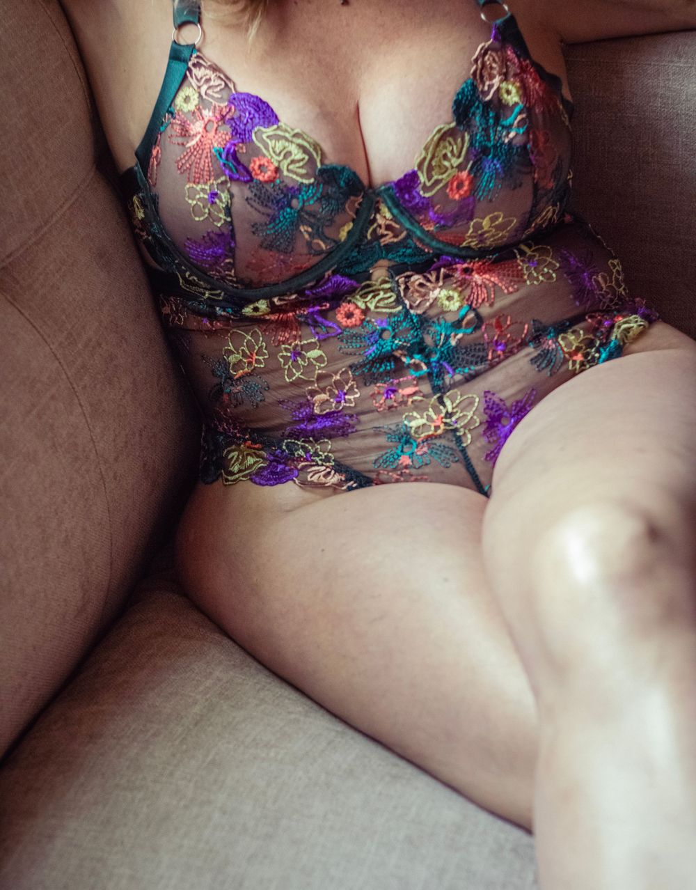 Woman wearing colorful floral embroidered lingerie bodysuit seated on a beige sofa.