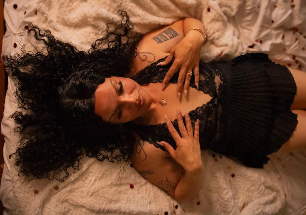 A person with curly dark hair lies on white bedding while posing artistically in warm lighting.