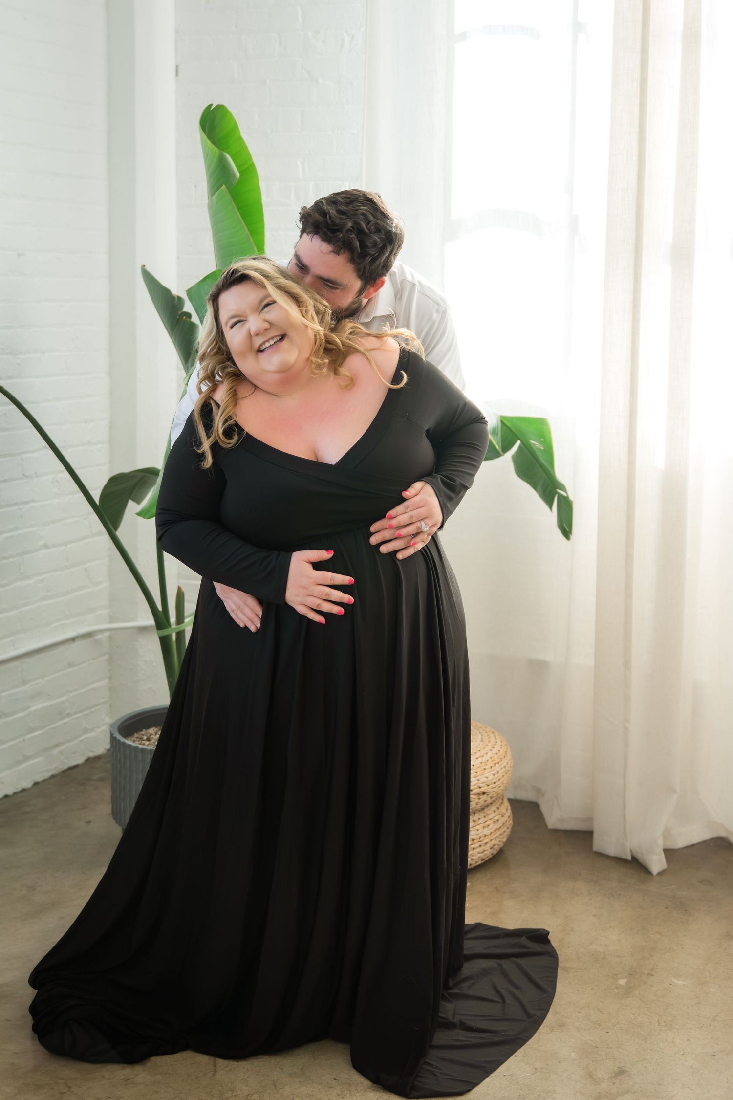 A couple shares an intimate moment in a minimalist studio setting with a black flowing dress and potted plant decor.