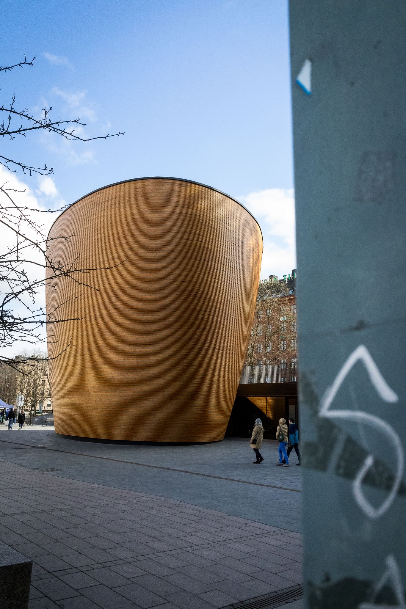 Golden copper cylindrical chapel building with people walking past on sunny day in city plaza.