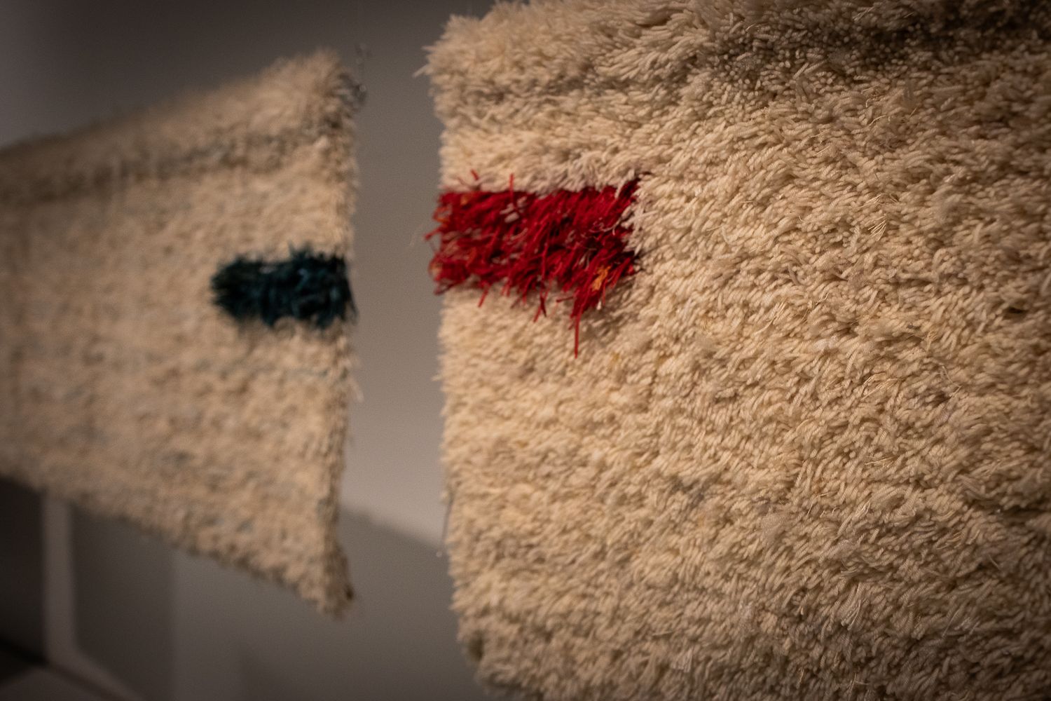 Textured cream wool wall hanging with small red and teal accent patches displayed in art gallery setting.