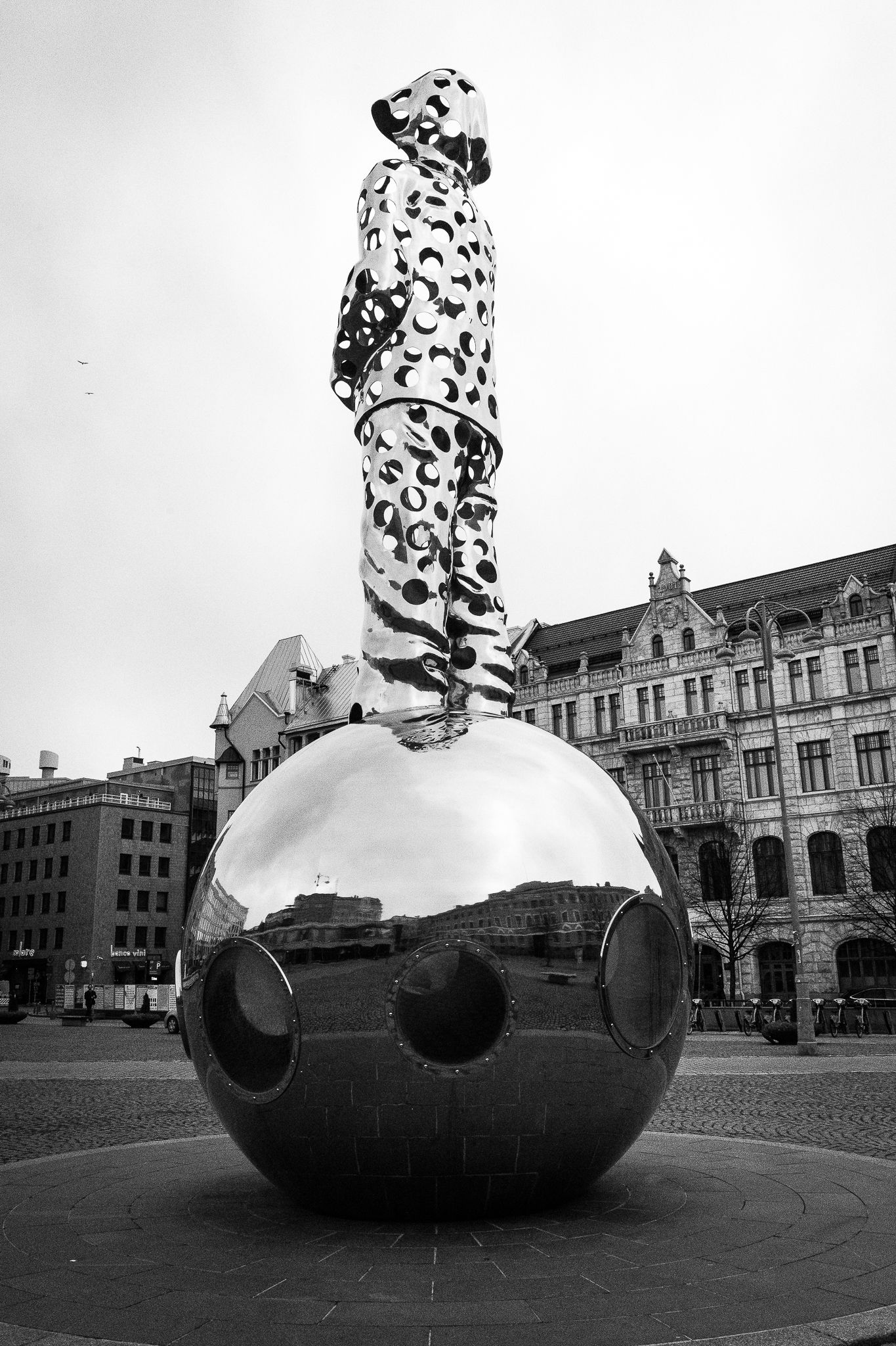 Black and white sculpture of figure in spotted suit standing atop large mirrored sphere in city square.