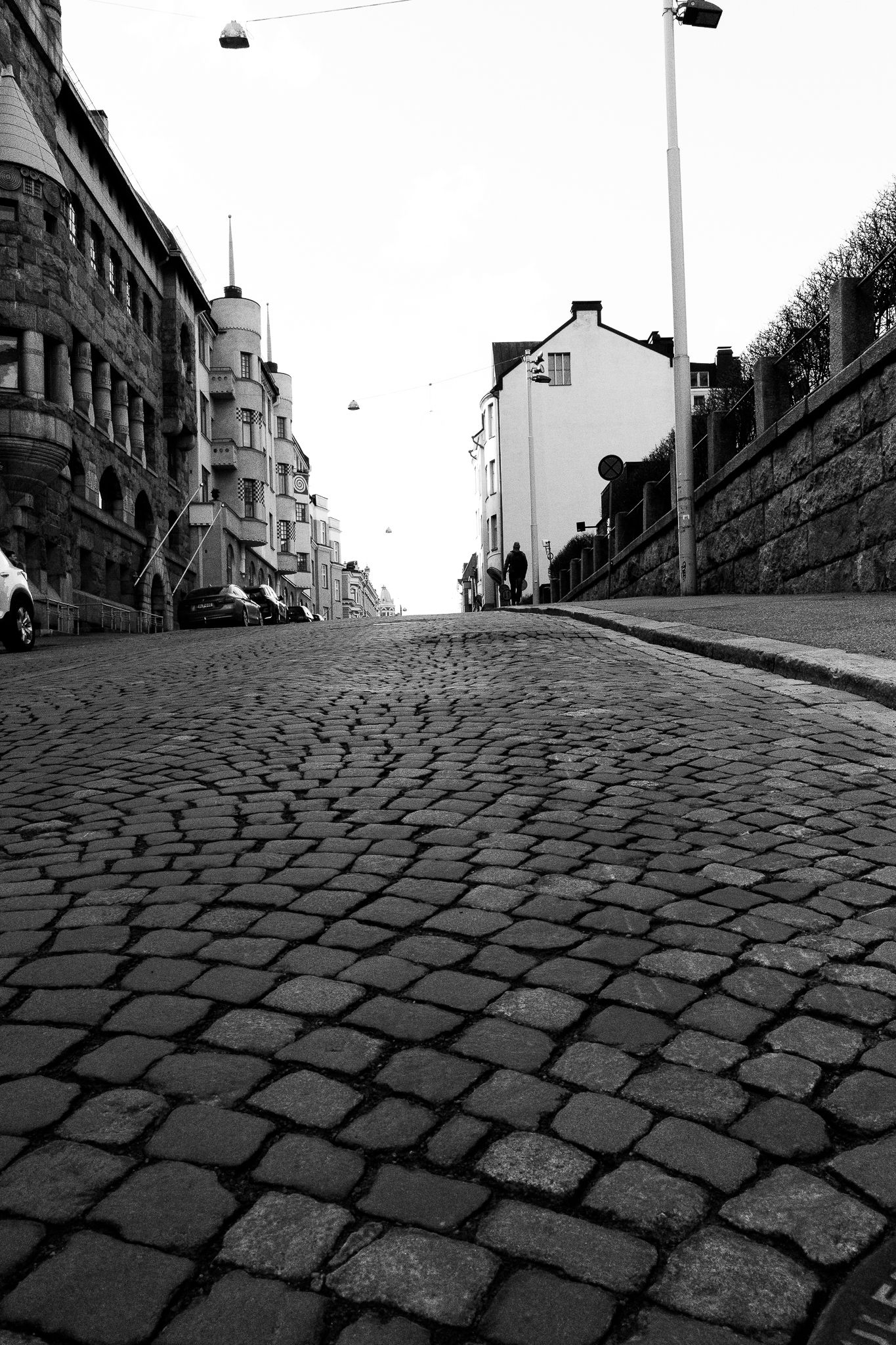 Black and white low-angle view of historic cobblestone street lined with buildings and lamp posts in European city.