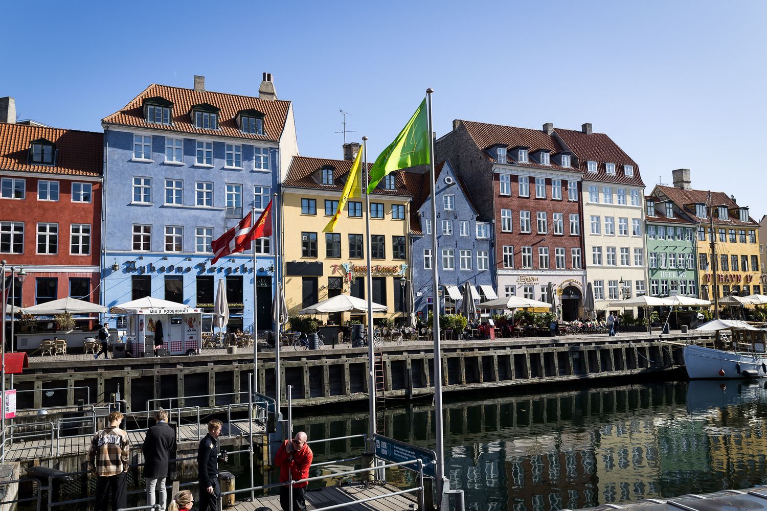 Tourists strolling along Nyhavn canal in Copenhagen with vibrant historic facades and international flags displayed.