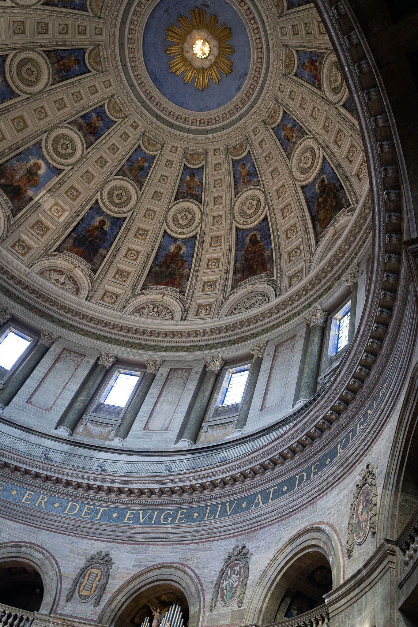 Interior view of an ornate cathedral dome with gilded details, painted frescos, arched windows, and Latin inscription.