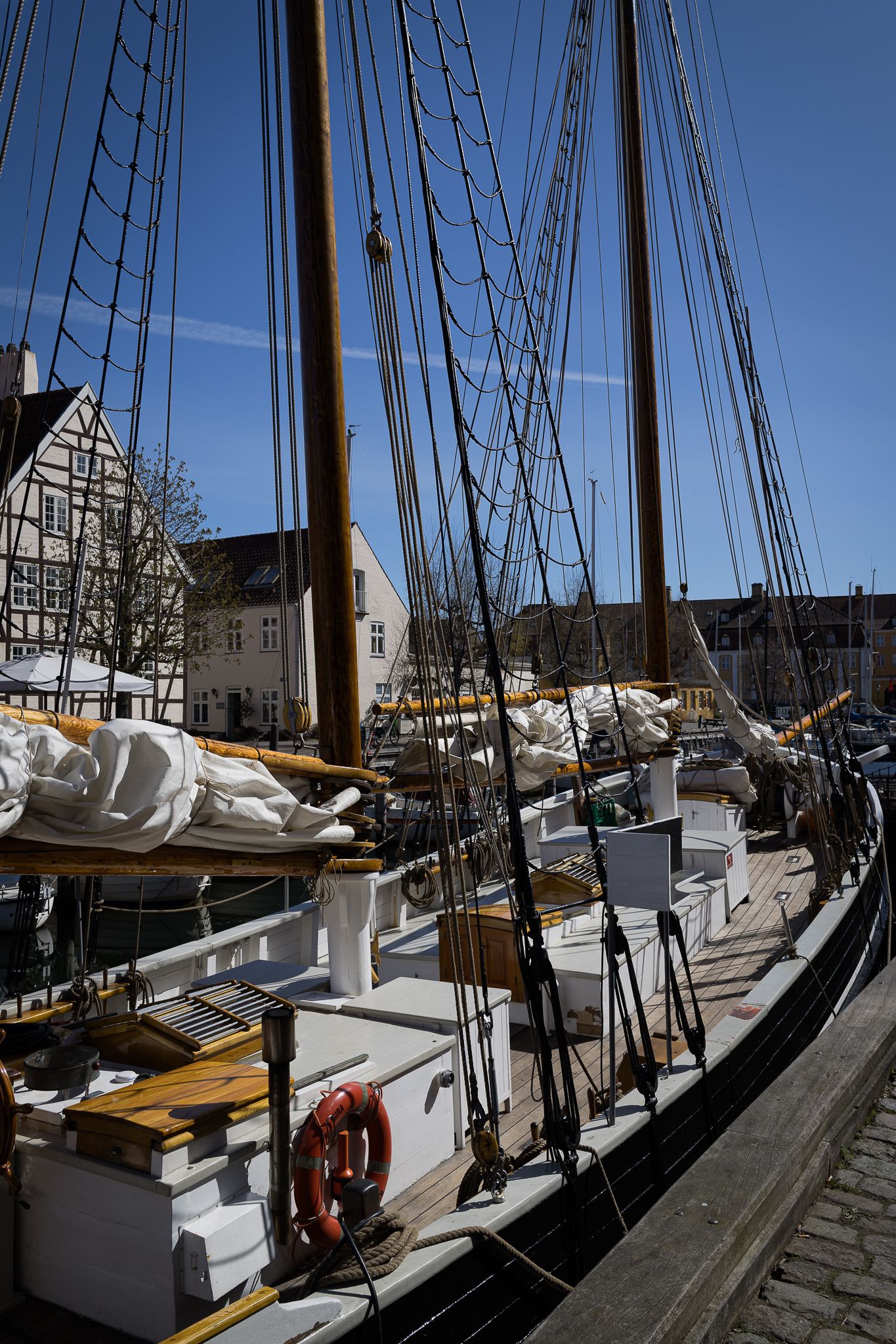 Vintage wooden sailing ship docked at a historic European harbor with traditional half-timbered buildings.