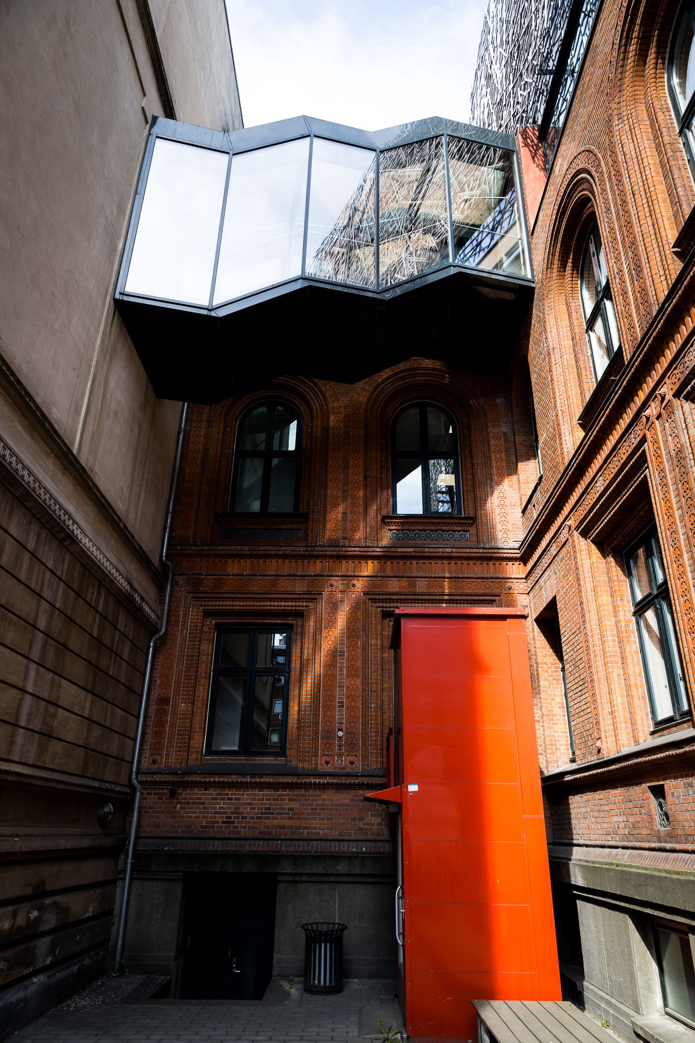 Historic red brick building with modern geometric glass extension and bright orange door in urban alley.