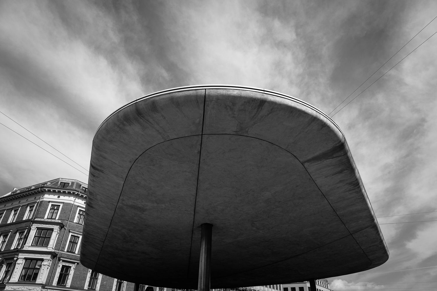 Black and white photo of a large curved concrete canopy structure against dramatic cloudy sky.