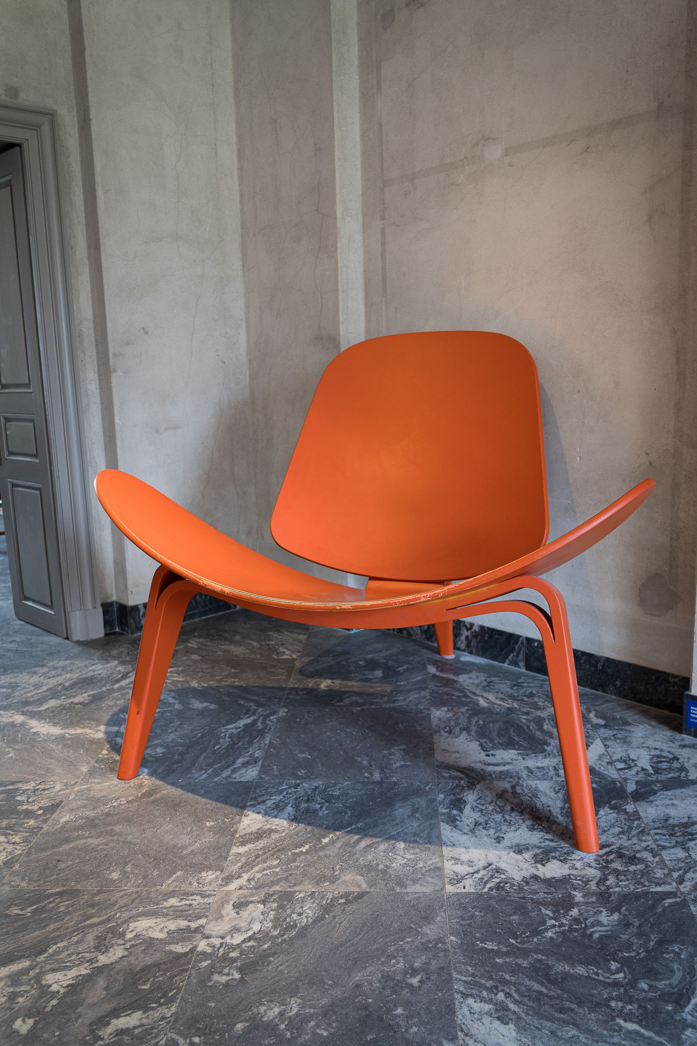 Iconic orange Hans Wegner Shell Chair on marble floor against concrete wall in minimalist interior.