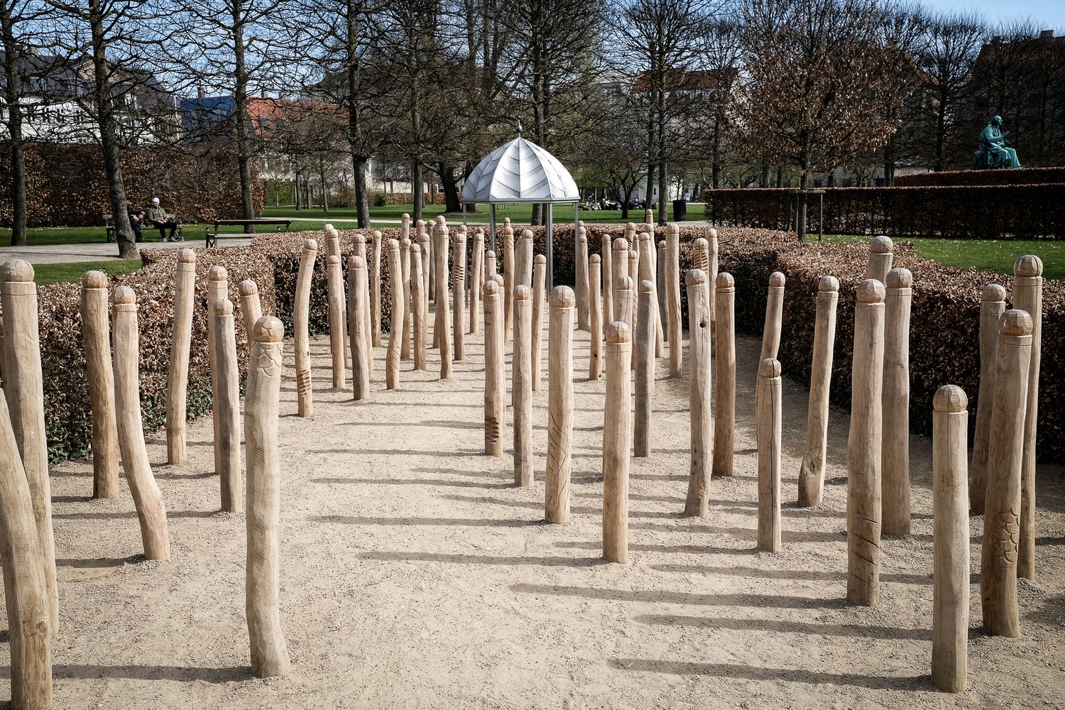 Rows of wooden log posts arranged in grid pattern on sandy ground in urban park setting.