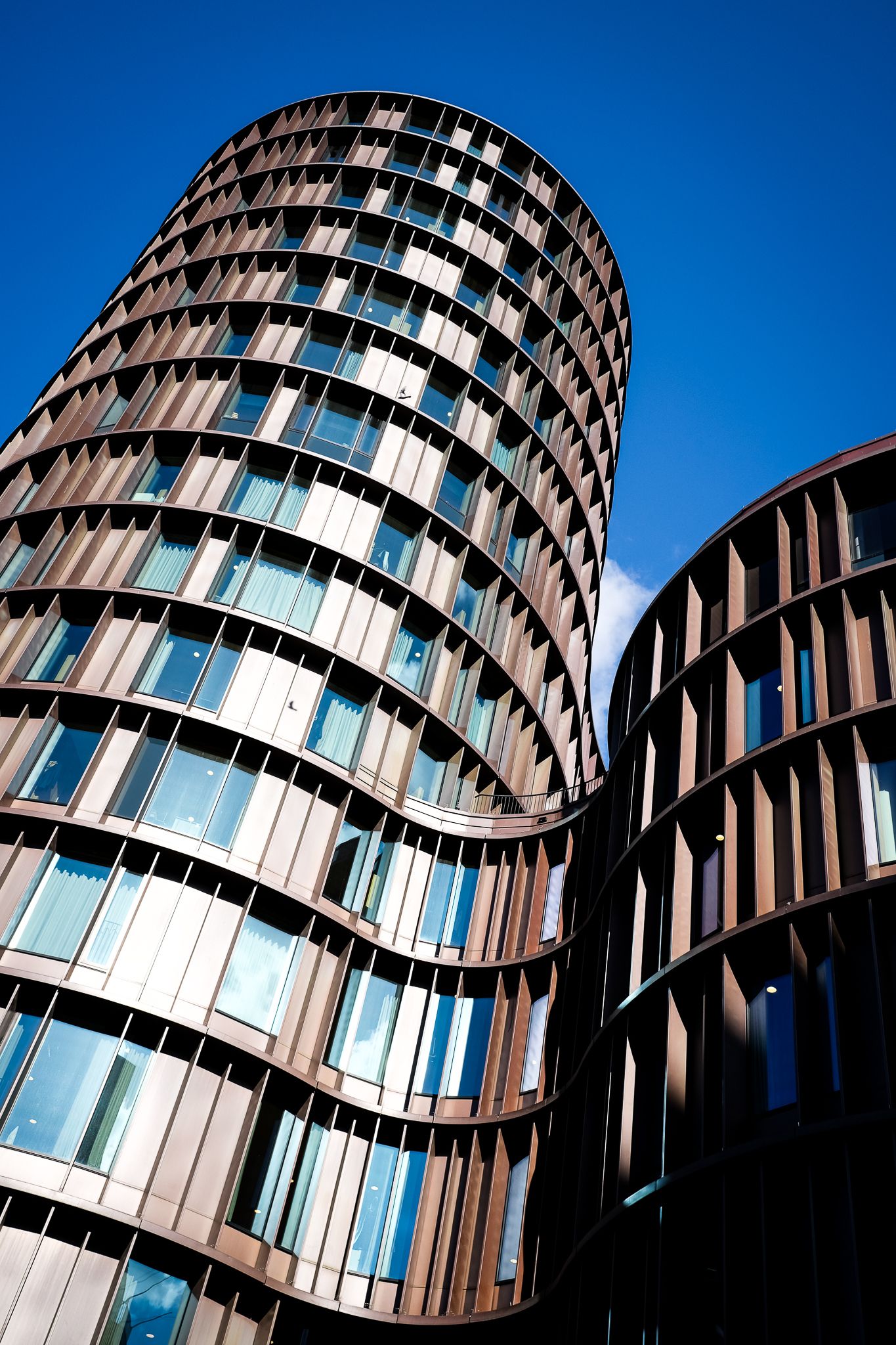 Modern curved cylindrical office towers with bronze metal facade and glass windows against blue sky.
