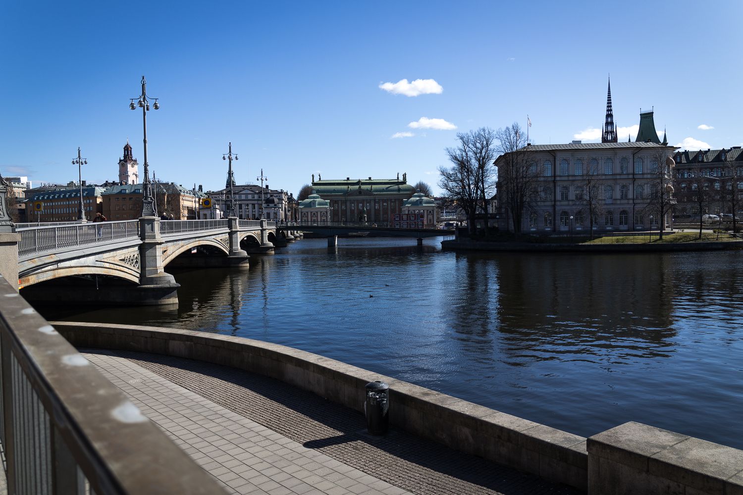 Scenic urban waterway with historic stone bridge, classical buildings and church spire under clear blue sky.