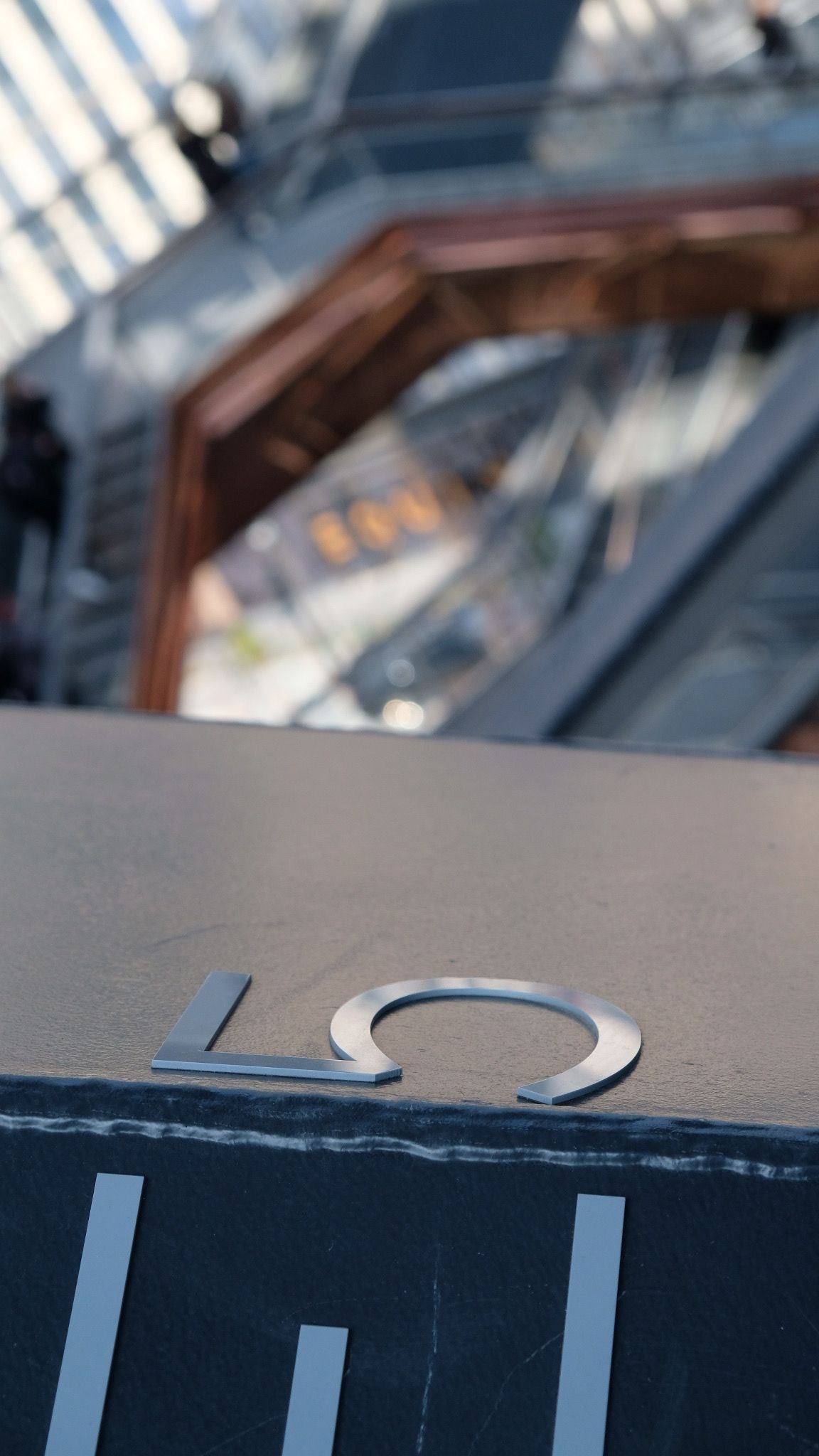 Close-up of a metallic floor number sign on a railing with blurred escalators in the background.