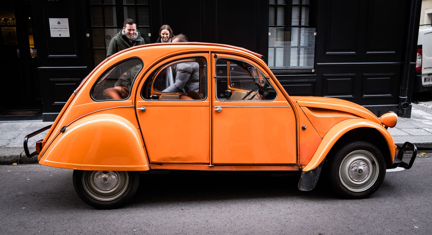 Vintage orange Citroën 2CV parked on a cobblestone street with two people visible in the background.