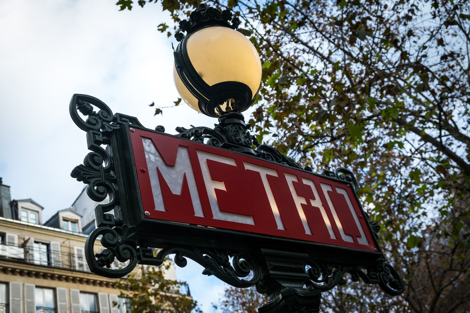 Classic ornate red Paris Metro sign on a decorative iron post with trees behind.