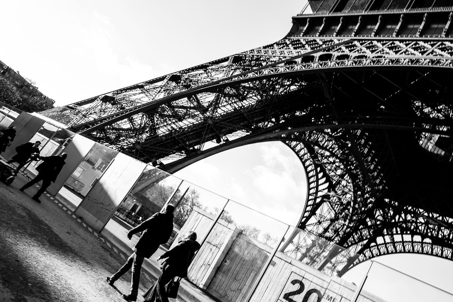 Black and white low-angle view of the Eiffel Tower base with pedestrians walking nearby.