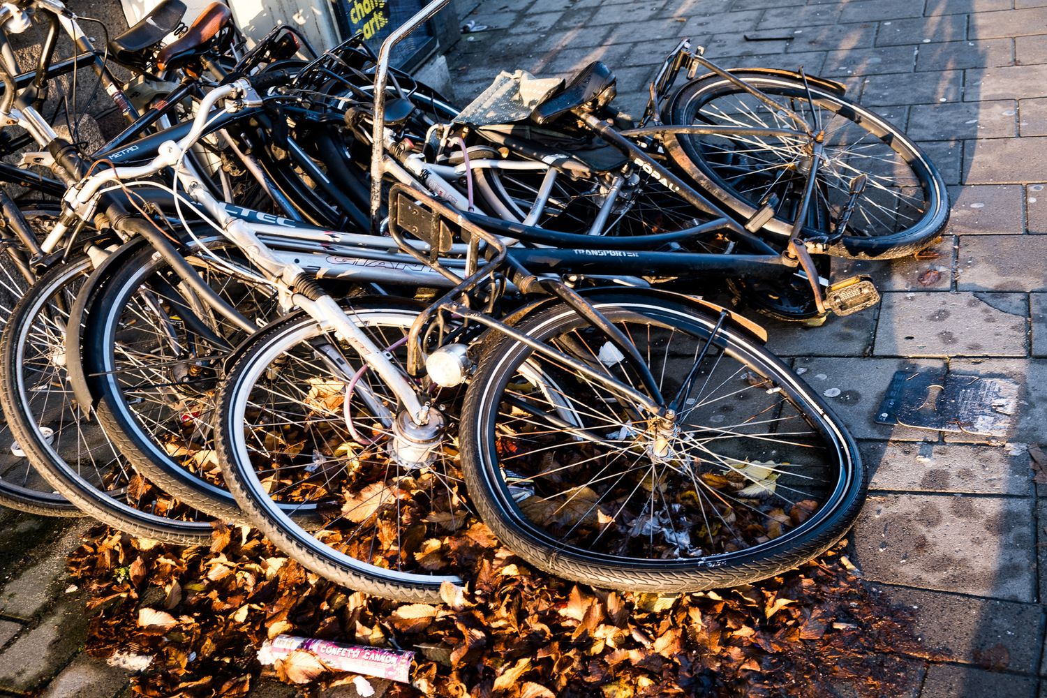 Pile of bicycles covered in autumn fallen leaves on cobblestone pavement.