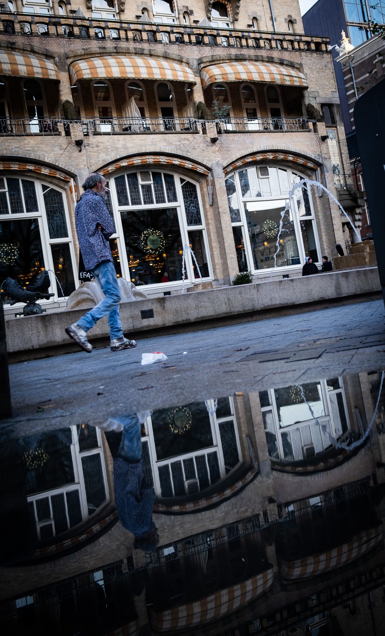 A pedestrian walks past a classic building reflected in a rain puddle on a city street.