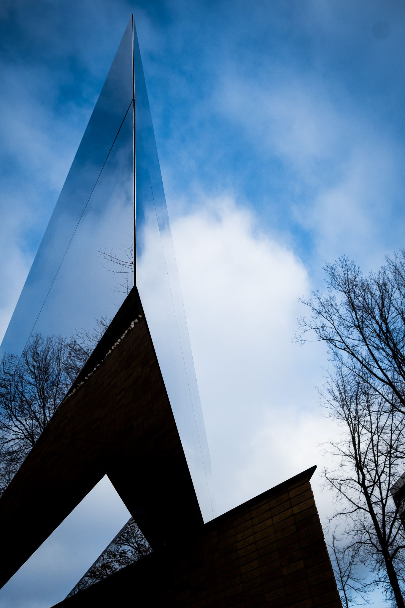 Dramatic low-angle view of a tall mirrored steel sculpture pointing toward a blue sky.