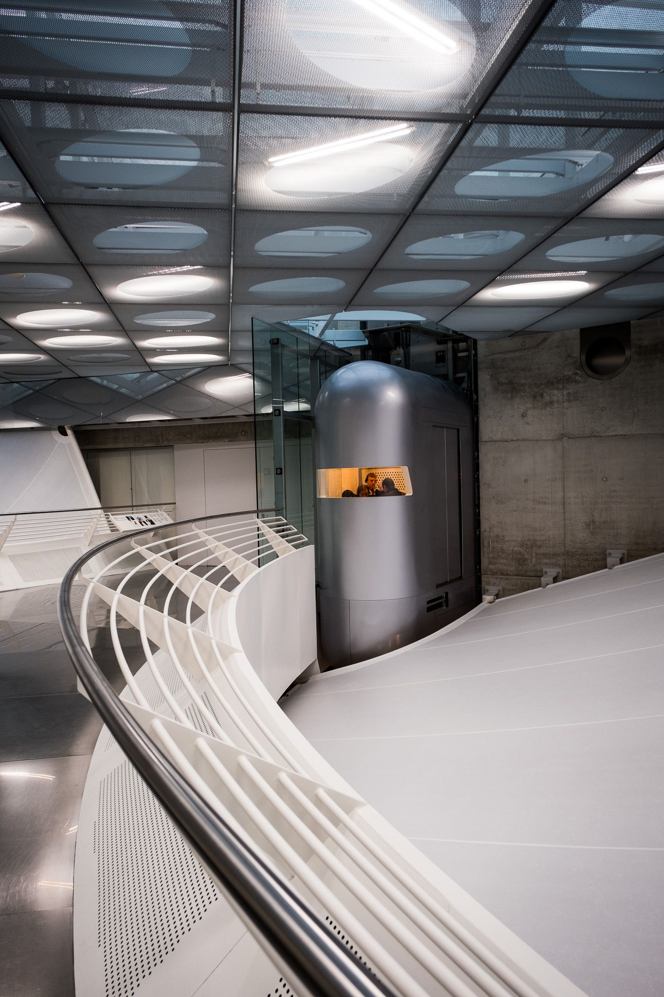 Futuristic curved interior corridor with glass ceiling panels, white railings, and a silver cylindrical elevator pod.