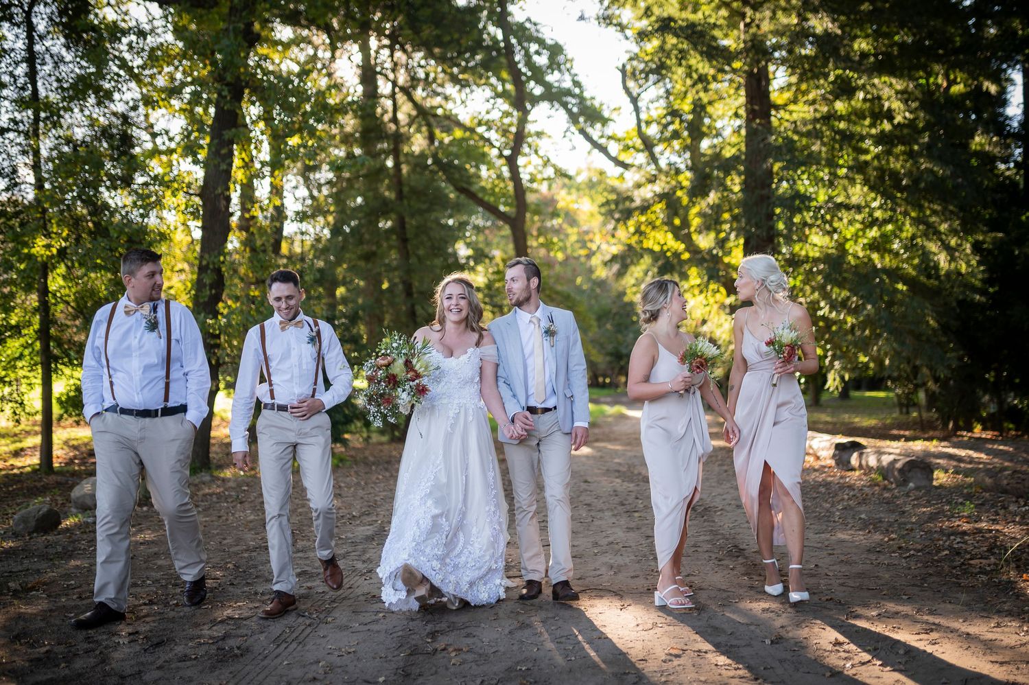 Wedding party walks outdoors among tall trees, bride in white gown with groomsmen in khaki and bridesmaids in blush pink.