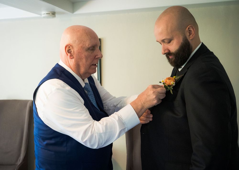 An older man pins a boutonniere on a bearded groom's lapel during wedding preparations.