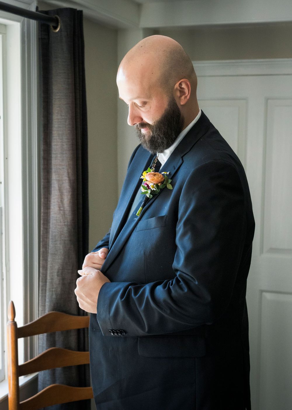 Bald groom with beard adjusting cufflinks in navy suit with floral boutonniere before wedding ceremony.