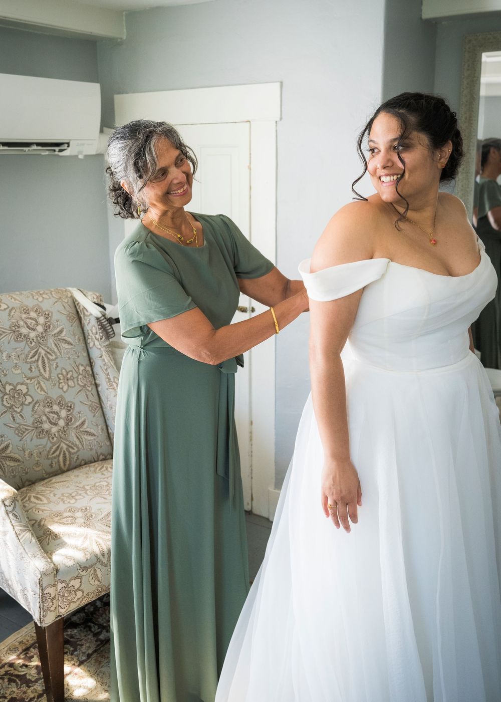 Bride smiling as older woman in green dress helps adjust her white off-shoulder wedding gown indoors.