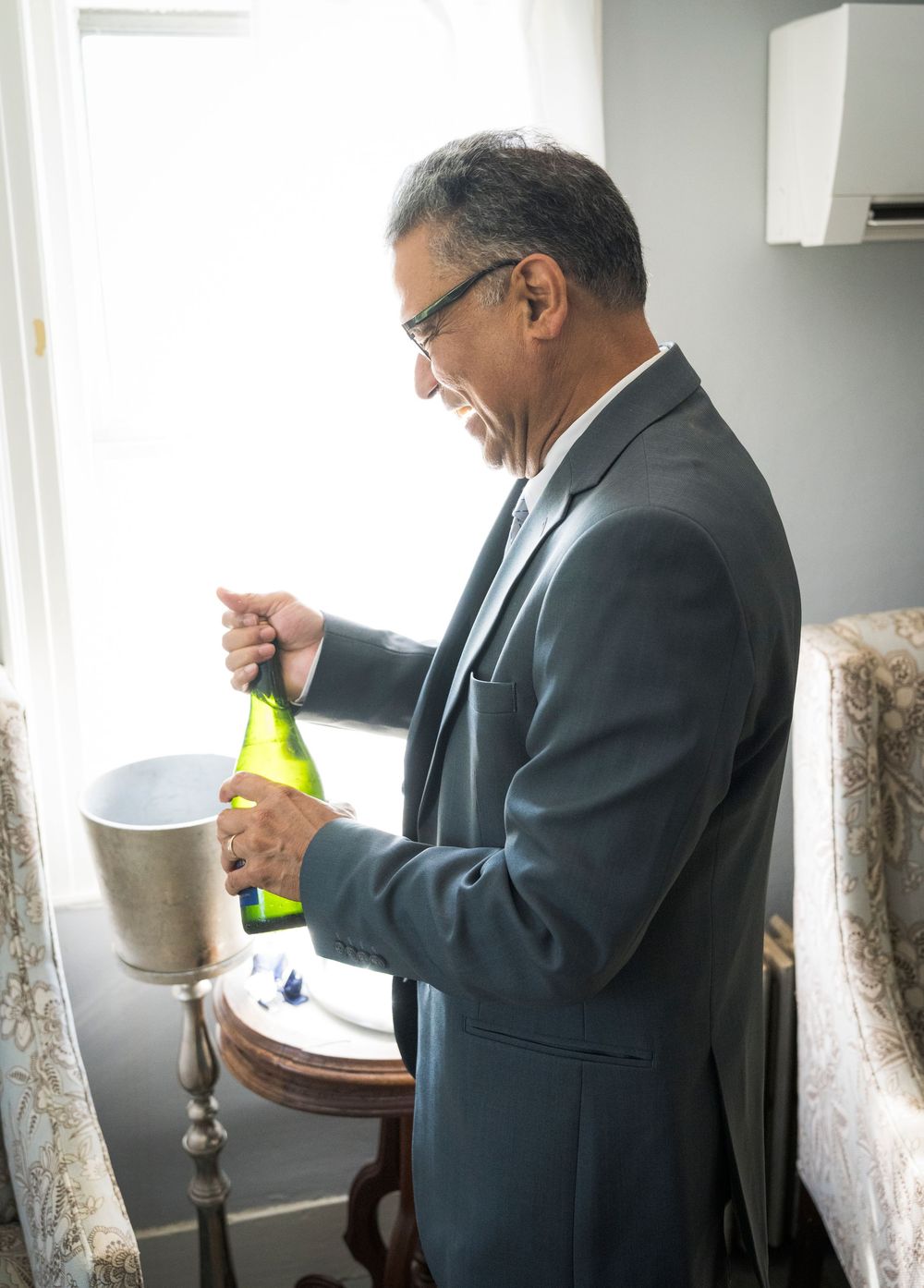 Man in suit opening a champagne bottle near a window with an ice bucket at a wedding reception.